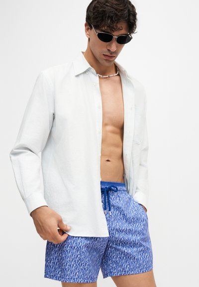 Modèle masculin portant des lunettes de soleil noires, une chemise blanche à manches longues ouverte, un collier de perles et un short de bain bleu à motifs, main dans la poche.