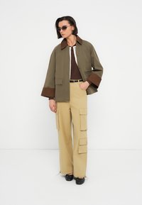 Χακί σακάκι με καφέ μανσέτες, συνδυασμένο με μπεζ παντελόνι cargo με πλαϊνές τσέπες. Το μοντέλο φοράει καφέ πικέ τοπ και γυαλιά ηλίου.