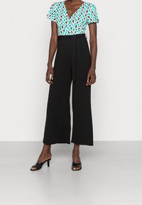 Barevně vzorované tričko s krátkým rukávem a černý overal s širokými nohavicemi a opaskem v pase. Černé sandály na vysokém podpatku s otevřenou špičkou dotvářejí vzhled.