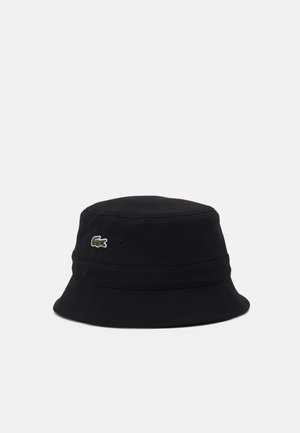 Chapeau seau noir en tissu texturé, présentant un logo Lacoste vert et blanc distinctif sur le côté. Couronne ronde, large bord.