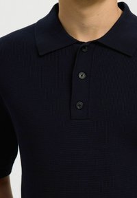 Polo bleu marine texturé avec trois boutons noirs et manches courtes porté par une personne.