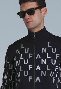Zwarte zip-up jas met een herhalend patroon van witte en grijze letters. Hoge kraag en een gestructureerde stof. Draag met donkere zonnebril.