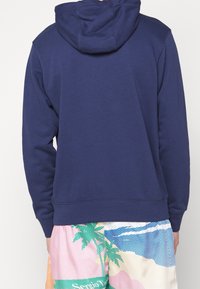 Mörkblå bomullströja med huva, utrustad med ribbad nederkant och ärmar, bärs med färgglada mönstrade shorts nedanför.