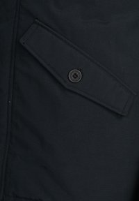 Tascone di giacca in tessuto nero con un ribattino rettangolare e chiusura a bottone rotondo nero, caratterizzato da un materiale testurizzato e cuciture pulite.