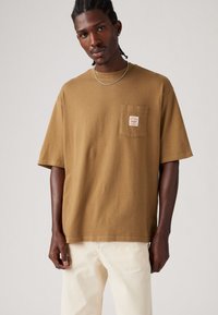 Tricou din bumbac maro, cu un buzunar pe piept și o etichetă. Croială lejeră, mâneci scurte și decolteu rotund. Asortat cu pantaloni de culoare deschisă.