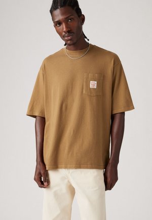 Tricou din bumbac maro, cu un buzunar pe piept și o etichetă. Croială lejeră, mâneci scurte și decolteu rotund. Asortat cu pantaloni de culoare deschisă.