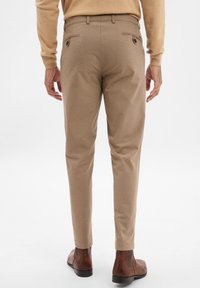 Beige tailored trousers with a slim fit, back pockets with flaps, and a smooth texture. Paired with brown ankle boots.