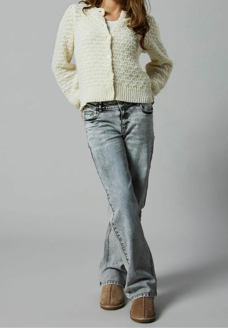 Femme portant un cardigan blanc texturé, un jean denim gris clair et des chaussures marron clair sans lacets, debout les mains dans le dos.