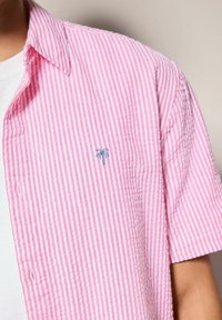 Kurzarmhemd mit einem rosa-weißen Streifenmuster, aus leichtem Stoff hergestellt, mit einer kleinen blauen Palme auf der Brust gestickt.