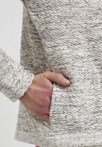 Pull gris clair texturé avec une finition tissée et des rayures sombres discrètes. Comprend une poche latérale et des poignets côtelés.