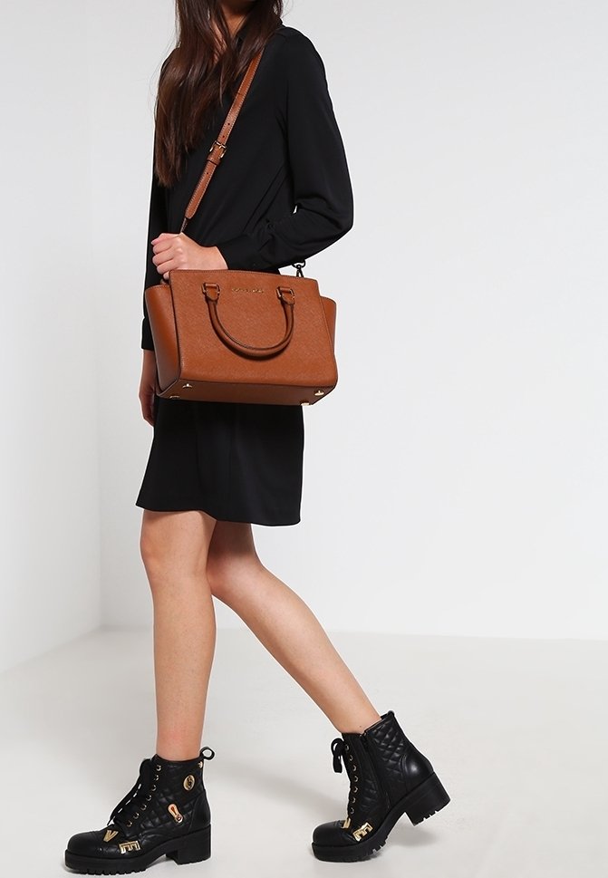 Femme vêtue d'une robe noire et de bottines noires à lacets, portant un sac à main en cuir marron avec des poignées et une bandoulière.