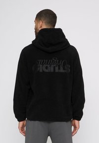 Mann mit kurzen Haaren, der eine schwarze Fleecejacke mit Kapuze trägt, auf deren Rückseite "STUDIO Originals" gedruckt ist, und von hinten vor einem weißen Hintergrund steht.