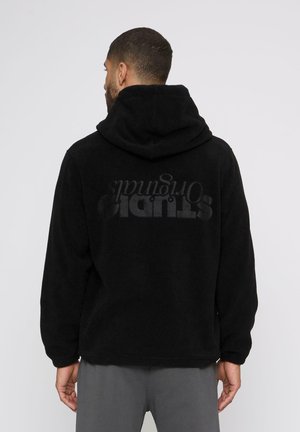 Mann med kort hår som har på seg en svart hettejakke i fleece med "STUDIO Originals" trykket på ryggen, vendt bort fra en hvit bakgrunn.