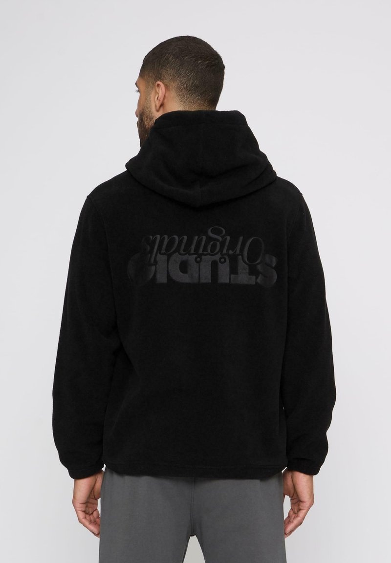 Mann mit kurzen Haaren, der eine schwarze Fleecejacke mit Kapuze trägt, auf deren Rückseite "STUDIO Originals" gedruckt ist, und von hinten vor einem weißen Hintergrund steht.