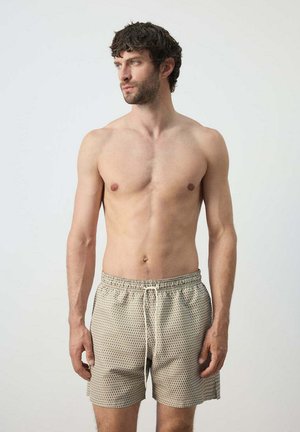 Hombre con cabello rizado y corto, vistiendo bañadores beige con estampado, de pie sin camiseta contra un fondo claro y liso, mirando hacia un lado.