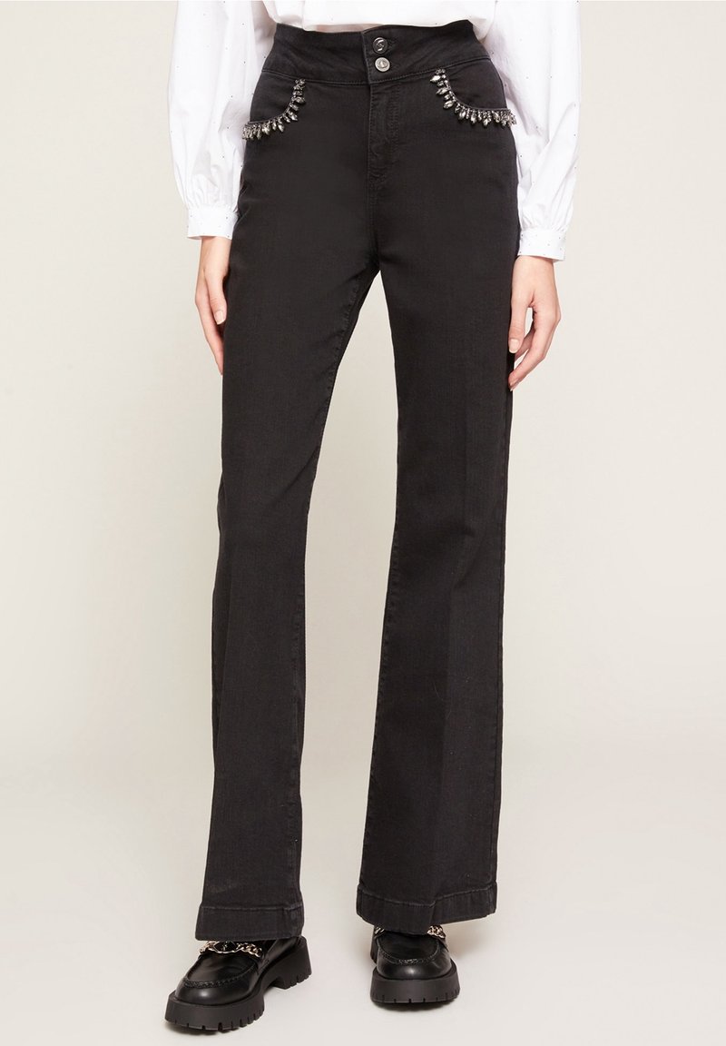 Femme portant un jean noir taille haute avec des strass décoratifs sur les poches avant, associé à des mocassins noirs épais et une blouse blanche à manches longues.
