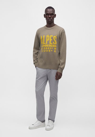 Homme portant un pull beige avec texte jaune "ALPES", pantalon gris et baskets blanches, debout devant un fond clair unicolore.