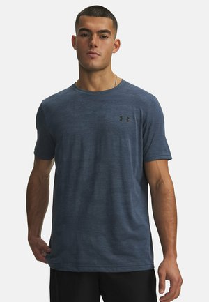 Jonge man met kort haar die een donkerblauw Under Armour T-shirt en zwarte shorts draagt, staand tegen een effen lichte achtergrond.