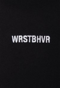 Tela negra con bordado en relieve blanco que dice "WRSTBHVR", con una textura suave y un ligero estiramiento.