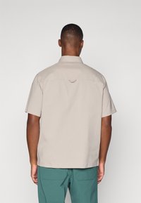Short-sleeve shirt in beige fabric, featuring a back center loop detail and a relaxed fit. Paired with green pants.