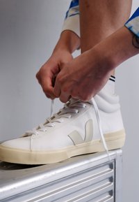 Bijele kožne tenisice s kremasto žutom gumom, s istaknutim V logom u svijetlosivoj boji i bijelim vezicama. Ruke vežu vezice.