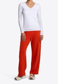 Weißes langärmliges V-Ausschnitt-Oberteil kombiniert mit weit geschnittenen orangen Hosen. Das Outfit wird mit klobigen Sneakern mit mehrfarbigen Akzenten abgerundet.