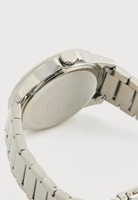 Silver metal wristwatch with linked bracelet, visible back case engraved with brand and material details, and a ridged crown on the side.