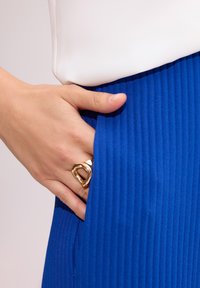 Niebieska spódnica z prążkowanego materiału, z ręką włożoną w kieszeń, akcentowana złotym pierścionkiem na palcu. Prosty biały top jest widoczny u góry.