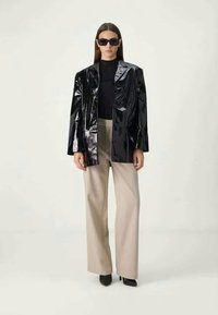 Mulher vestindo um blazer preto brilhante, top preto, calças largas bege, saltos pretos e óculos escuros escuros, de pé contra um fundo liso.