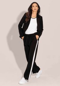 Zwarte op maat gemaakte blazer met een wit T-shirt en zwarte broek met witte zijsstripes. Het model draagt witte sneakers en toont een casual look.