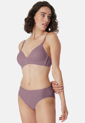 Femme aux cheveux bouclés et foncés portant un soutien-gorge et des sous-vêtements mauves assortis, regardant vers sa gauche avec un bras croisé sur sa taille.