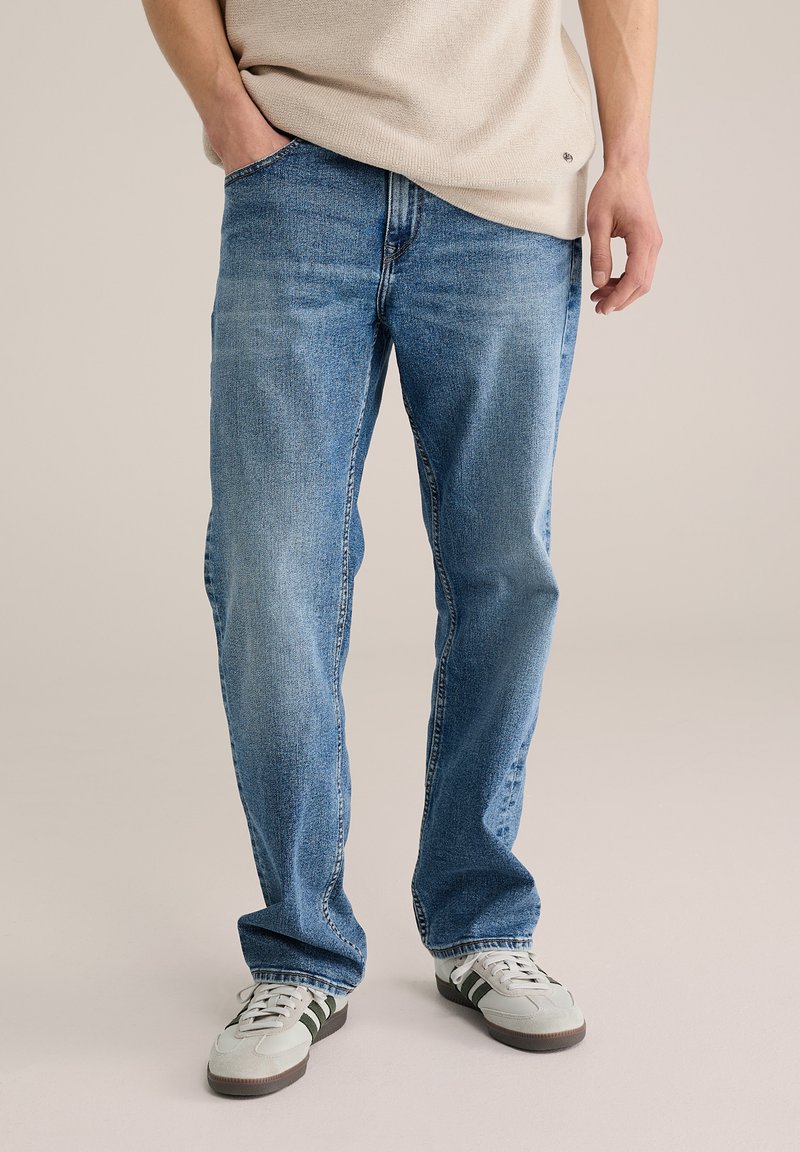 Šviesiai mėlynos džinsinės kelnės tiesiomis kojomis, turinčios tradicinį penkių kišenių dizainą ir subtilų išblukimą, derinamos su pilkais sportbačiais.