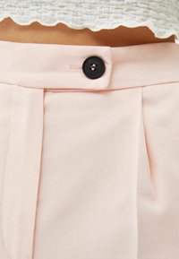Světle růžové kalhoty s texturovanou tkaninou, s předním skladem a černým knoflíkem na zapínání v pase. Jednoduchý a přizpůsobený design.