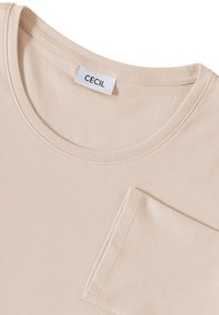 Beige bomull t-shirt med rund halsringning, korta ärmar och en ficka på vänster sida; etiketten säger "CECIL." Slät textur.