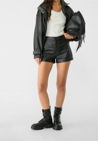 Zwarte neplederen jas met drukknoopjes, witte getailleerde tanktop, zwarte leren short en zwarte veterschoenen. Inclusief fringetas.