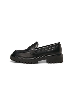 Mocasín de cuero negro con suela gruesa de taco, costura de mocasín y una correa sobre la parte superior, mostrado en perfil lateral sobre fondo blanco.
