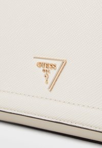 Kremfarget faux skinn med en teksturert overflate og en gull trekantet logo med "GUESS USA" og et spørsmålstegn.