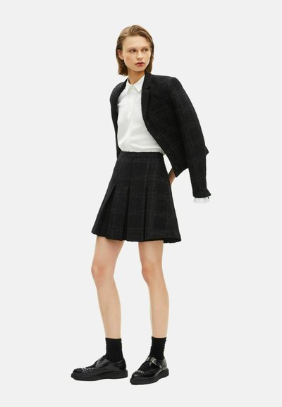 Zwarte tweed blazer en rok set, met een gestructureerde afwerking, boxy snit en plooien. Gecombineerd met een witte button-up shirt en zwarte schoenen.