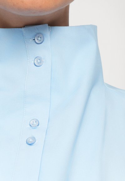 Light blue, collared shirt with a smooth texture, featuring four matching buttons along the placket and neat stitching details.