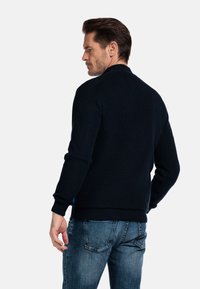 Marineblauer Strickpullover mit gerippter Textur und Stehkragen, kombiniert mit hellblauen Jeans. Die Bündchen und der Saum sind ebenfalls gerippt.