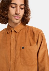 Koeienvacht overhemd in warm bruin met knoopsluiting aan de voorkant, kraag en een linker borstzak met een logo patch. Gelaagd stof met ribbelpatroon.