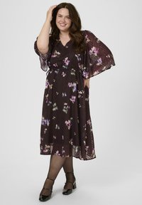 Sötét virágmintás ruha V nyakkivágással, széles ujjakkal és rugalmas derékrésszel. Lila és zöld virágminták találhatóak a texturált anyagon. Fekete magassarkú cipő.