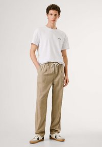 Weißes Baumwoll-T-Shirt mit kleinem Logo auf der Brust, kombiniert mit beigen Jogginghosen mit Kordelzug und weißen Sneakers mit schwarzen und roten Akzenten.