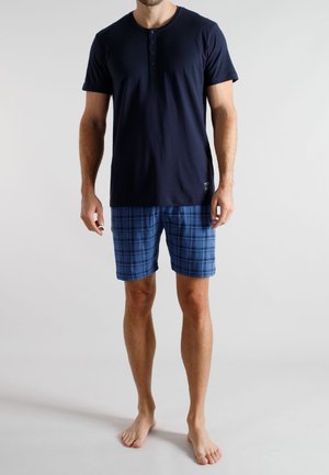 Mann steht barfuß und trägt ein marineblaues Henley-Shirt mit kurzen Ärmeln sowie blaue karierte Shorts vor einem schlichten hellen Hintergrund.