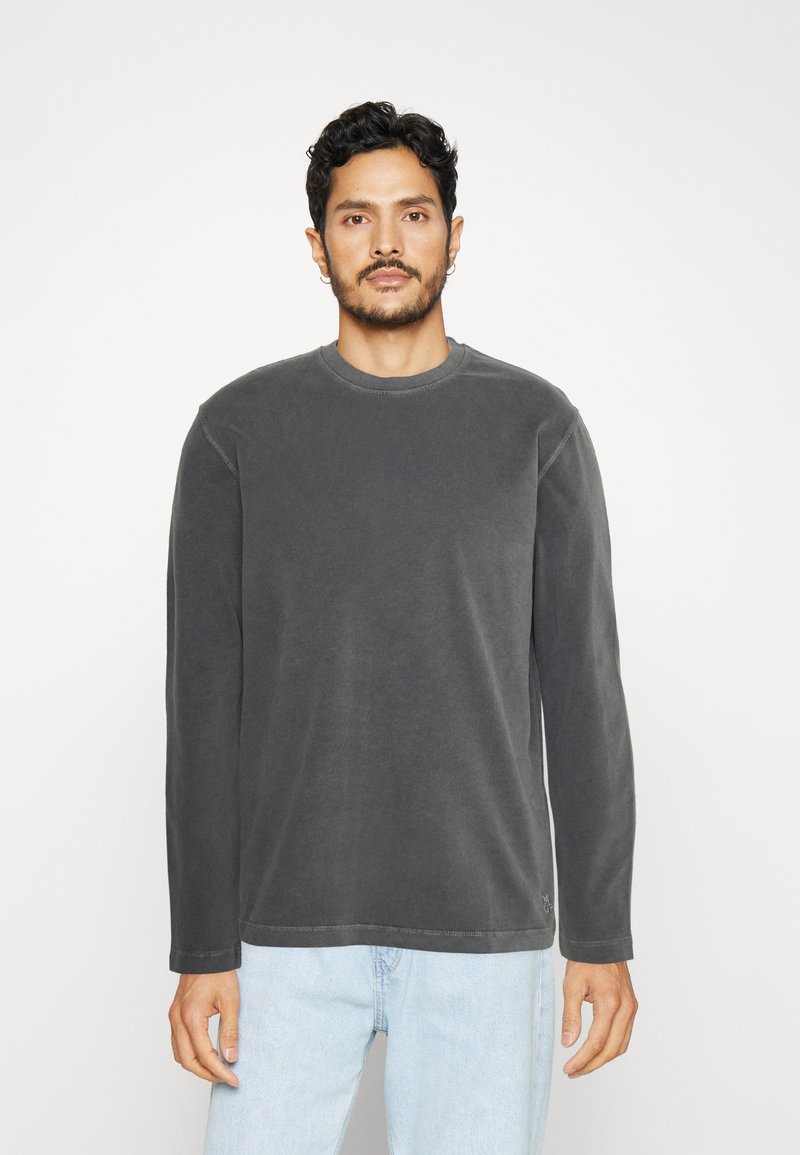 Camisa de manga larga con cuello redondo en gris oscuro. Tejido suave con una textura lisa y un sutil detalle de costura. Combinada con jeans de mezclilla azul claro.