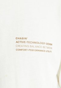 Bijela tkanina s ispisanim tekstom u svijetlosmeđoj boji: "CHASIN' ACTIVE-TECHNOLOGY-DENIM STVARANJE RAVNOTEŽE IZMEĐU UDOBNOSTI-PERFORMANSI-KORISTI."