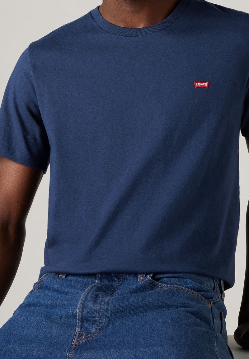 Personne portant un T-shirt Levi's bleu foncé uni avec un petit logo rouge Levi's sur la poitrine et un jean en denim bleu.