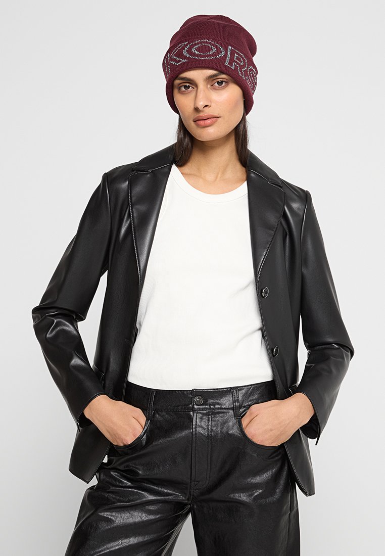 Schwarze Kunstleder-Blazer mit Reverskragen, weißem Rundhalsausschnitt-T-Shirt und bordeauxroter Strickmütze mit auffälligem Logo-Text. Schwarze glänzende Hose.