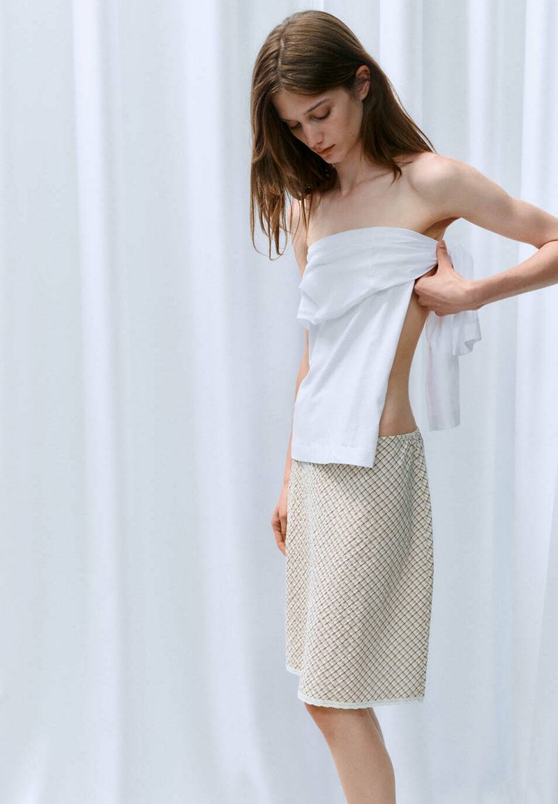 Jeune femme ajuste un top blanc sans bretelles tout en portant une jupe à motifs longueur genoux sur fond blanc.