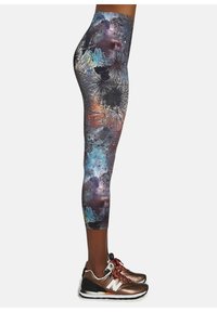 Legginsy w wielokolorowy nadruk galaktyczny z wysokim stanem, w odcieniach fioletu, niebieskiego oraz skomplikowanych czarnych wzorach. Noszone z brązowymi sneakersami.